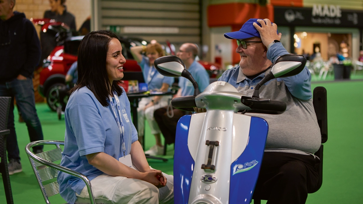 A female staff member sat down talking to a male powered scooter user at Motability Scheme Live