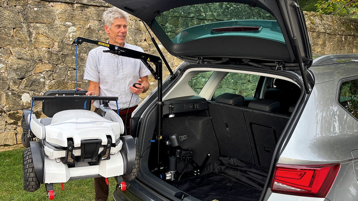 An elderly male driver, stood beside the open boot of his vehicle, operating the Swift Lift scooter hoist.