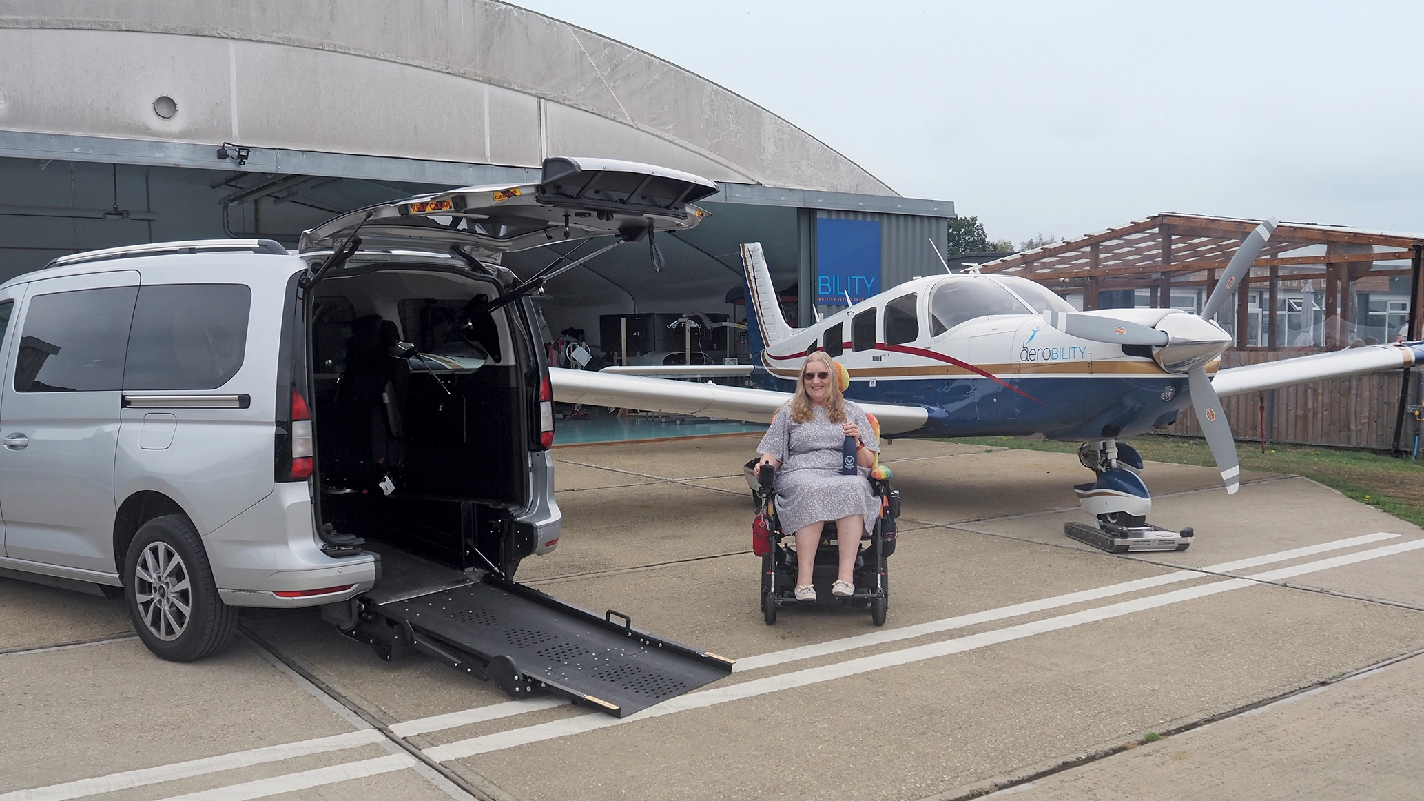 A young female wheelchair used pictured between a WAV and a light aircraft