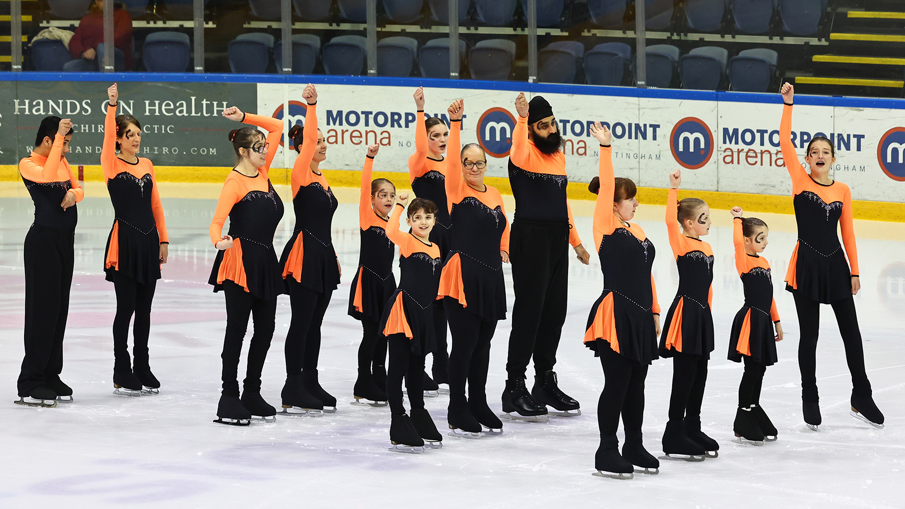 Ice-skaters dressed in orange and black and lifting one arm up
