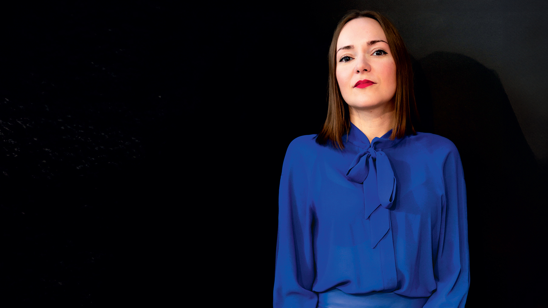 Victoria Jenkins wearing a blue shirt and red lipstick in front of a black background 