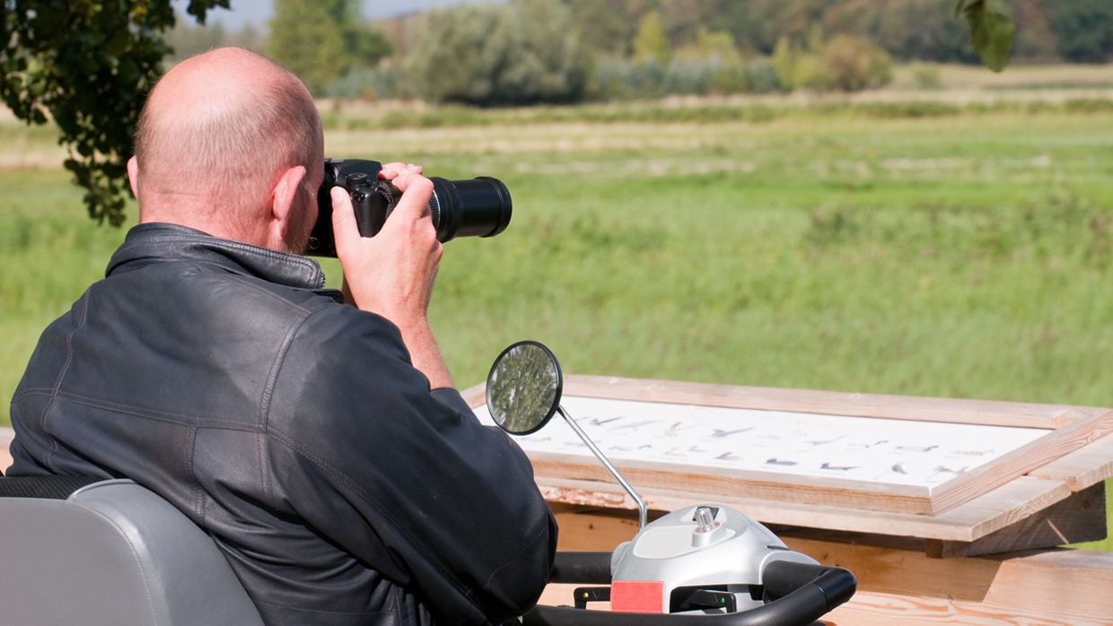A wheelchair user birdwatching with a camera, sat on a decking looking out over a fielded area