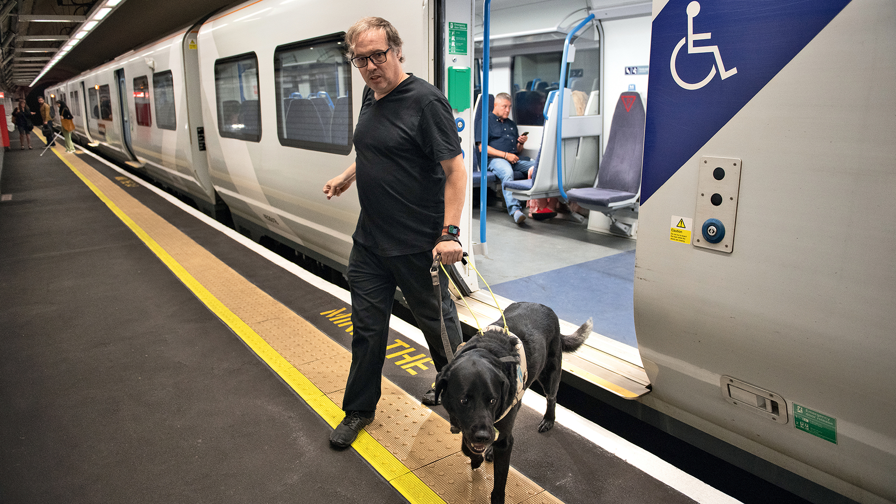 Man wearing glasses and a black t-shirt and trousers with a guide dog coming out the tube