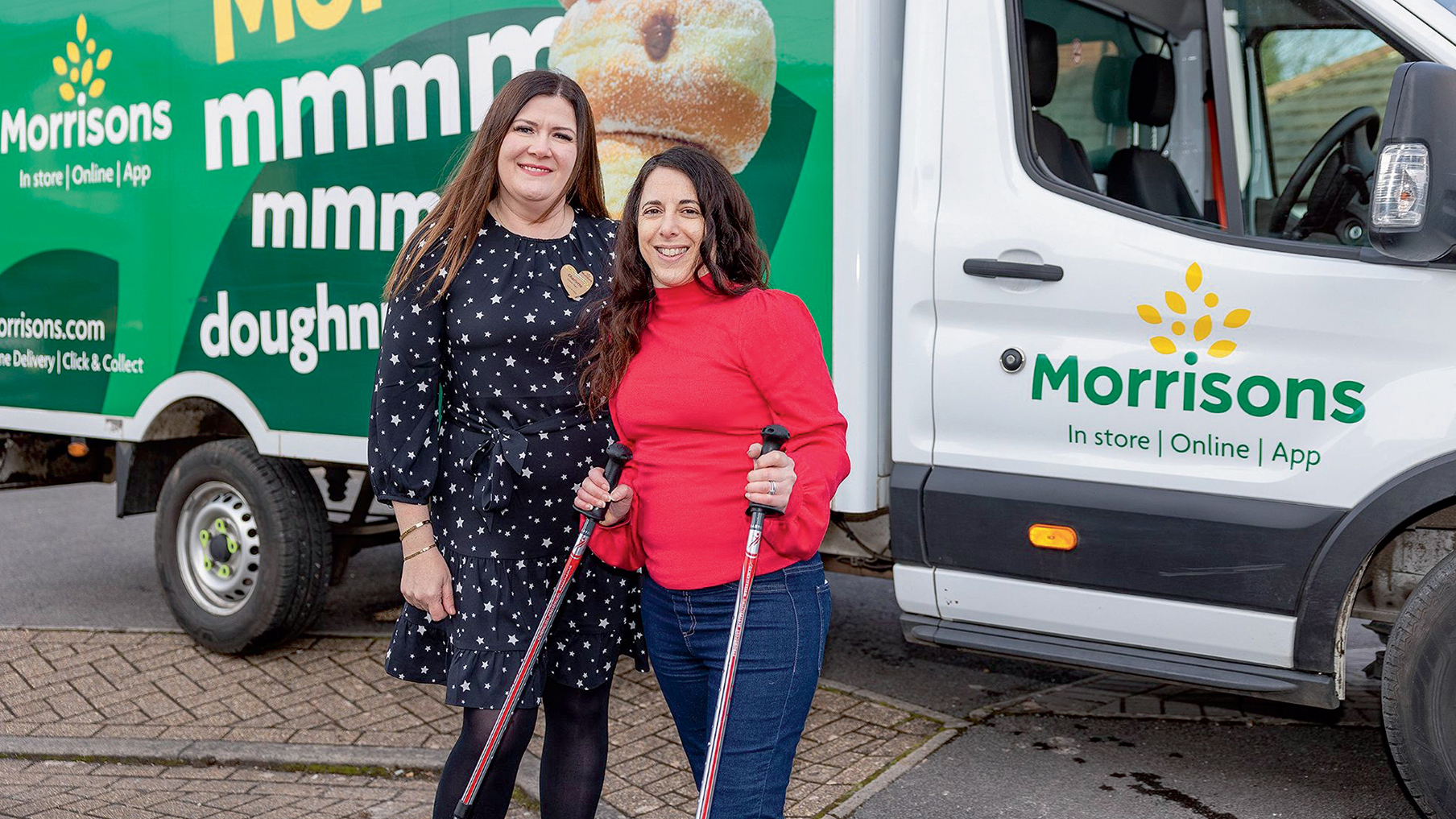 two women, one wearing a black dress and another a red jumper, are smiling at the camera, holding their walking sticks